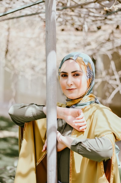 A long-sleeved shirt in yellow, wearing a blue and white scarf, leaning on the grey metal column of women
